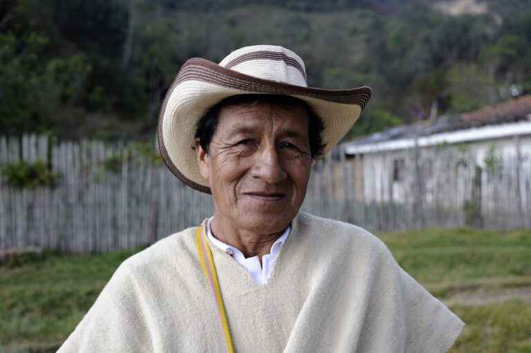 An older man in Colombia wears a brown and cream hat as he looks into the camera. He wears a beige poncho on his person with the strap of a yellow and pink bag showing. He stands in front of a fence with greenery around him.