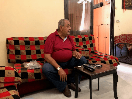 Ibrahim Aoun, an older Lebanese man, sits on a red, black, and brown checkered couch. He wears a red polo shirt as he looks off to the side.