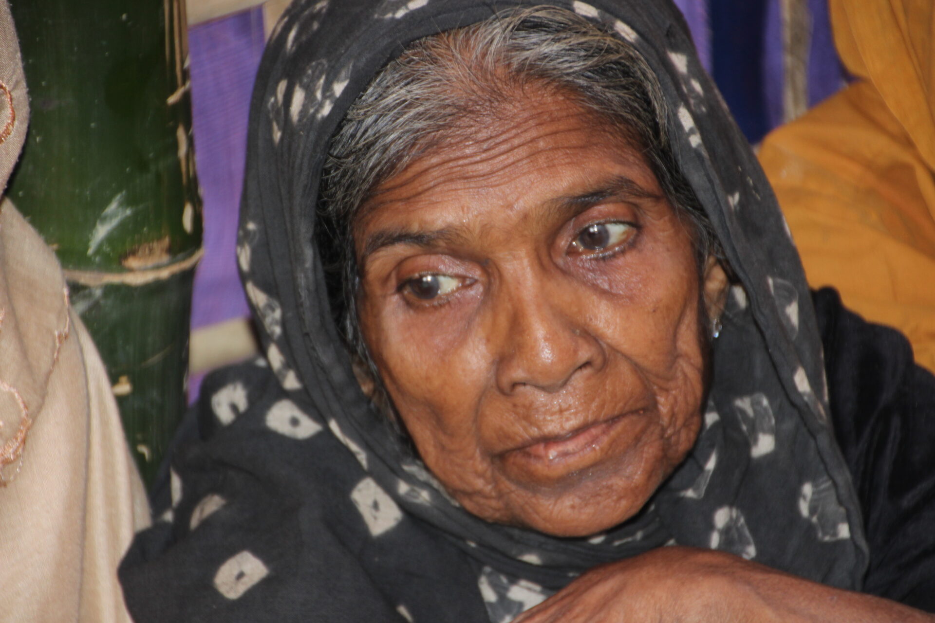 An older Rohingya woman in a headscarf looks down.