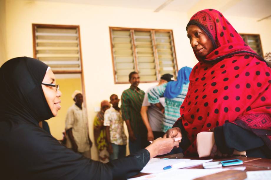 A woman in a black hijab and glasses smiles as she greets a woman in a red and black polka dot hijab. A line forms behind the woman in the red and black polka dot hijab.