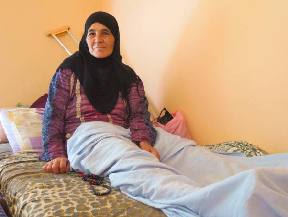 Syrian woman lays in bed as she recieves healthcare. She is wearing a black hijab and a crutch lays behind her. A white blanket covers her legs.