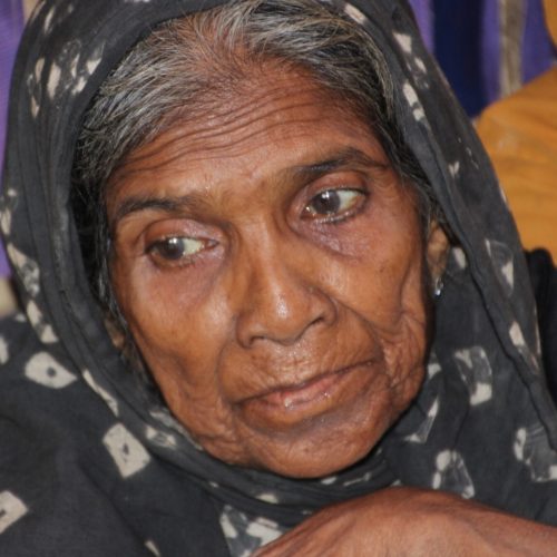 An older Rohingya woman in a headscarf looks down.